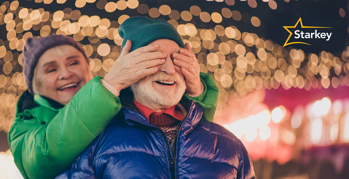 Image of senior woman covering husband's eyes amidst holiday lights in the background